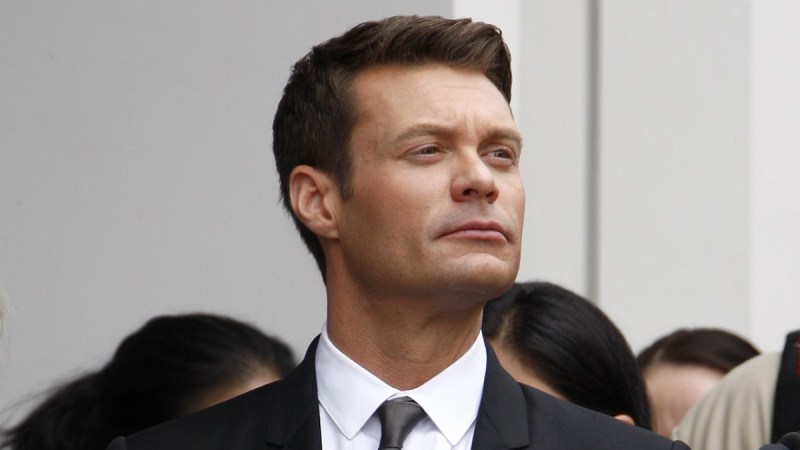 Ryan Seacrest wears a black suit and gazes out with a serious expression