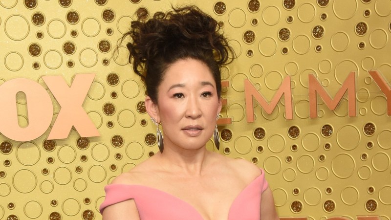 Sandra Oh wears a pink dress against a gold background on the red carpet