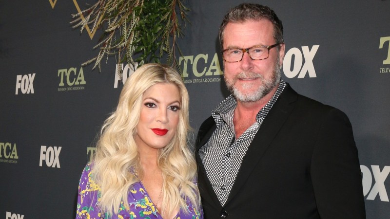 Tori Spelling, in a purple floral gown, stands with Dean McDermott, in a dark suit, on the red carpet