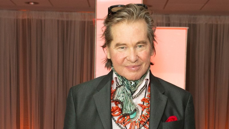 Val Kilmer wears a gray suit and brightly colored scarf around his throat