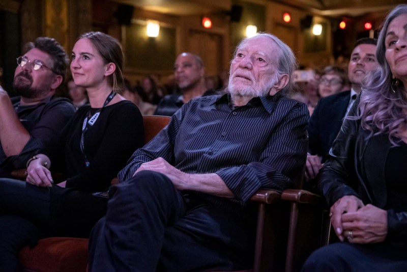 Willie Nelson in a black shirt and pants sitting in a theater audience