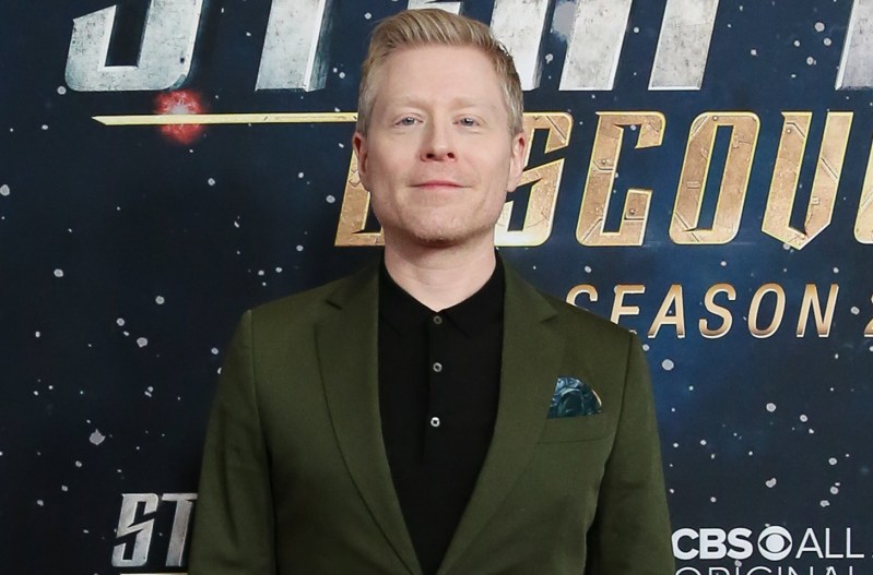 Anthony Rapp, standing at a red carpet even in a black shirt and green blazer