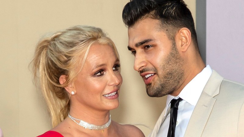 Britney Spears wears a red dress and stands with Sam Asghari, in a beige suit, on the red carpet