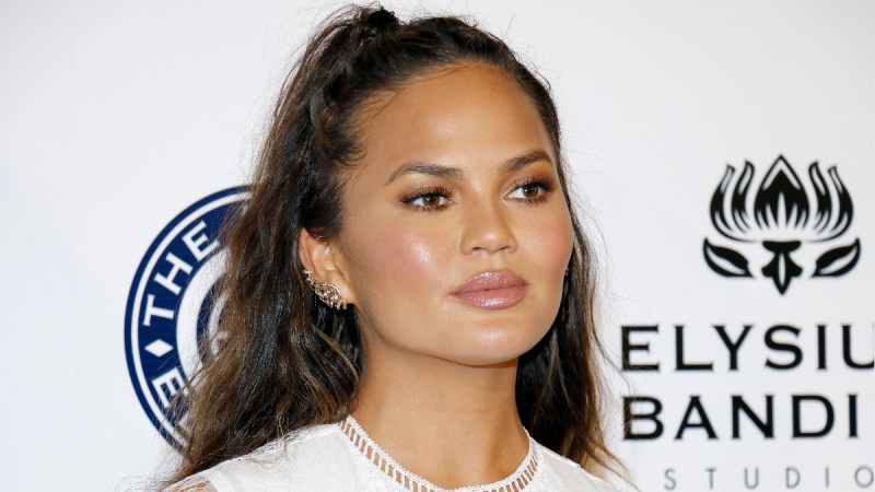 Chrissy Teigen wears a white dress on the red carpet and poses in front of a white background