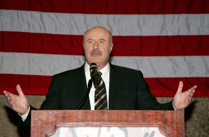 Dr. Phil with his arms out, standing at a podium, in front of an American Flag