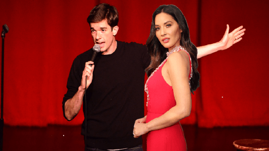 A photo of John Mulaney wearing a dark shirt performing onstage. Imposed over it is an image of Olivia Munn in a red dress