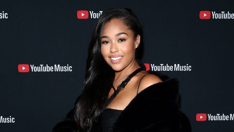 Jordyn Woods wears a black dress and coat against a black background on the red carpet