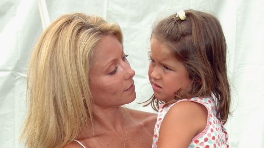 Kelly Ripa, in a tan top, holds daughter Lola Consuelos, in a white and red dress, in her arms outdoors
