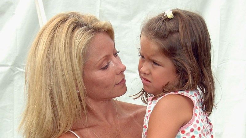 Kelly Ripa, in a tan top, holds daughter Lola Consuelos, in a white and red dress, in her arms outdoors