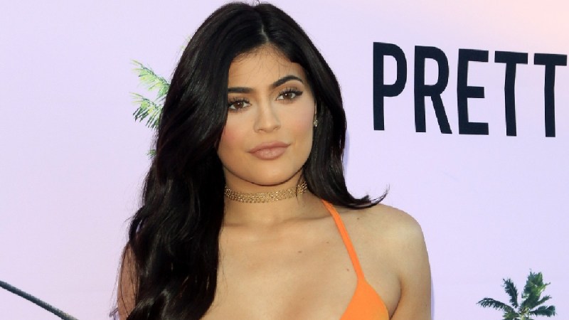 Kylie Jenner wears an orange dress against a pale pink background on the red carpet