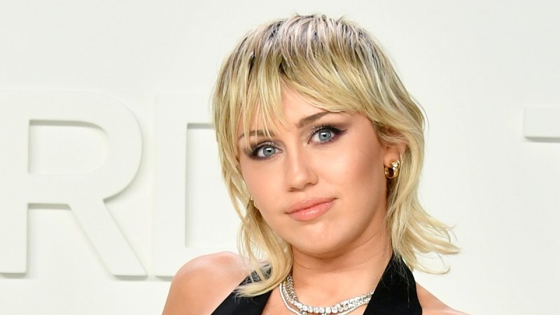 Miley Cyrus wears a black jumpsuit with a plunging neckline against a white background