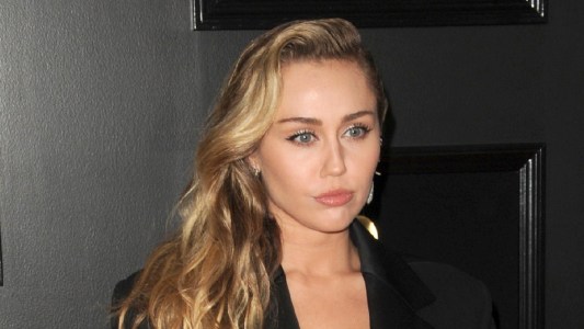 Miley Cyrus wears a black suit against a black background on the red carpet