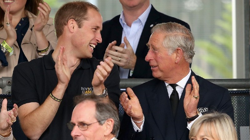 Prince William, in a black shirt, claps and smiles at Prince Charles, in a black suit, at the Invictus Games