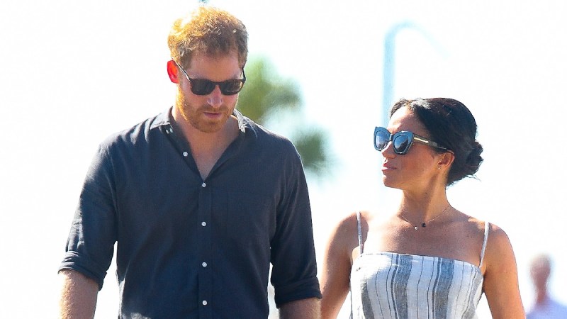 Prince Harry, in a dark shirt, walks outdoors with Meghan Markle, in a white dress