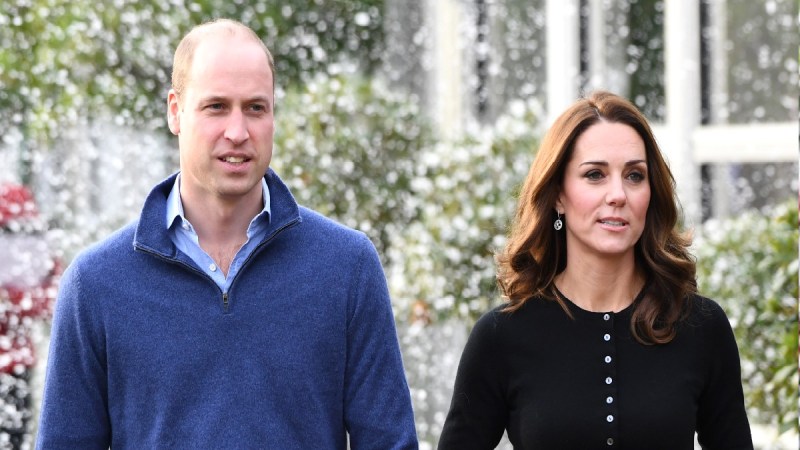 Prince William, in a blue sweater, walks with Kate Middleton, in a black top, outdoors
