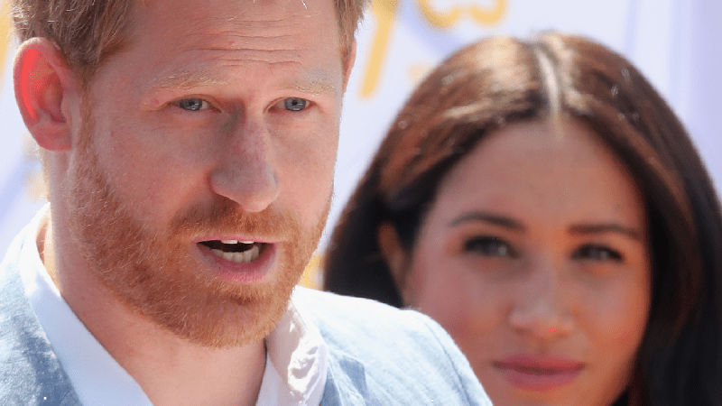 Prince Harry wears a blue suit as he gives a speech while Meghan Markle watches from behind