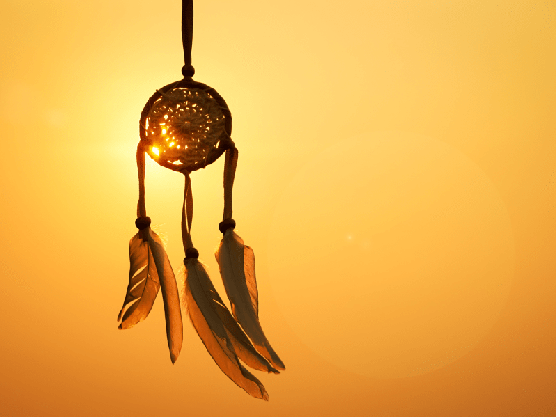 A dreamcatcher with large feathers hangs with a sunset in the background.