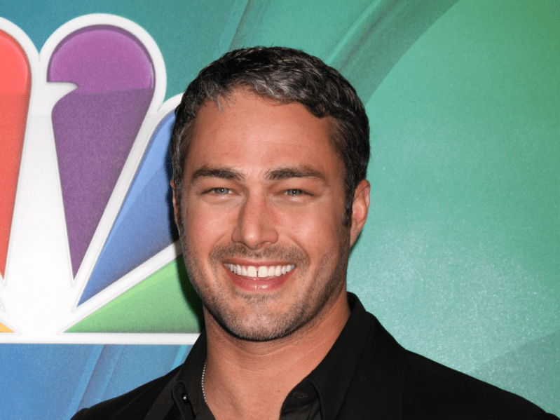 Taylor Kinney grins at the camera in a black button up shirt