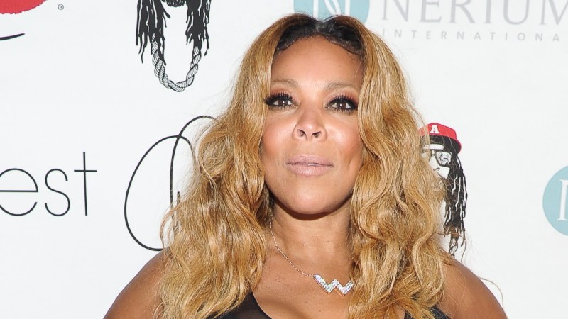 Wendy Williams poses in a black dress against a white background on the red carpet