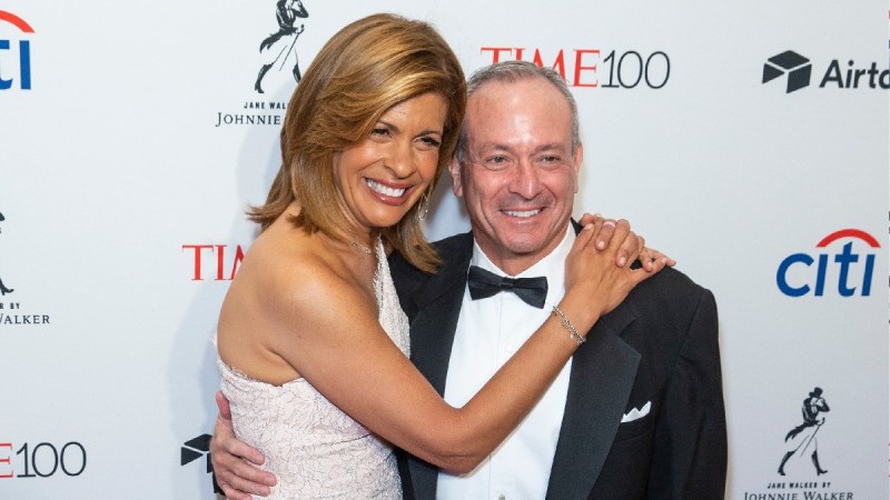 Hoda Kotb, in a white dress, stands with her arms around Joel Schiffman, in a black tux, on the red carpet