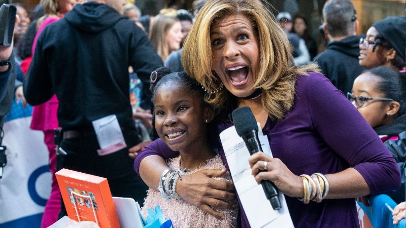 Hoda Kotb, in purple, cuddles up to a young girl while holding a microphone