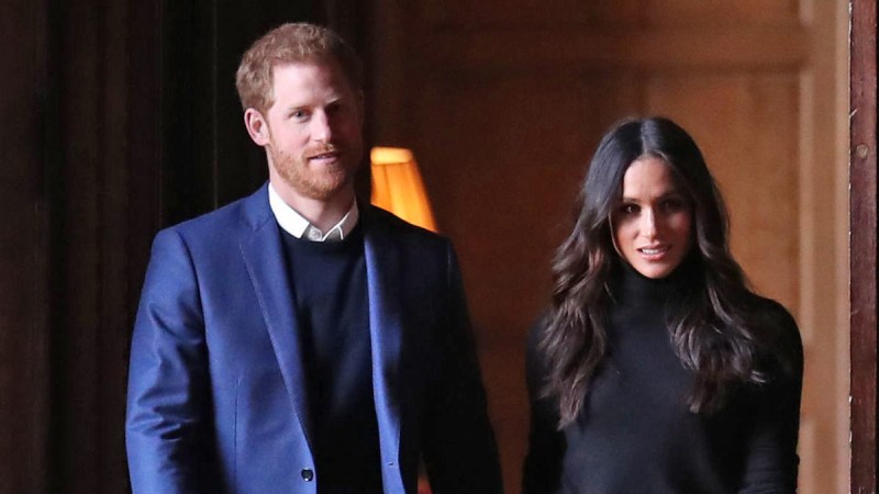 Prince Harry, in a blue suit jacket, walks with Meghan Markle, in a black top, down a hallway in Edinburgh, Scotland