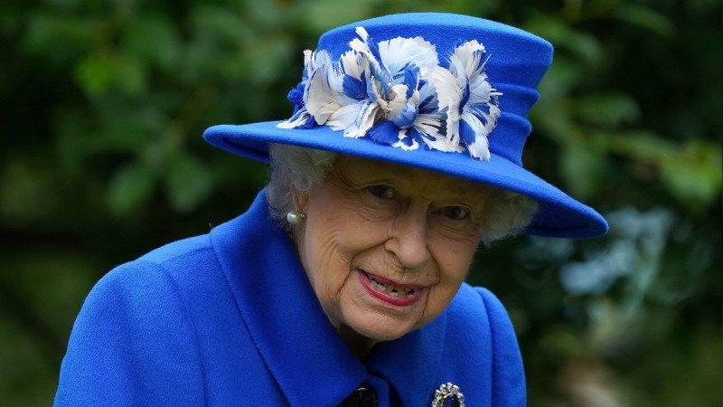 Queen Elizabeth wears a blue hat and coat outdoors against a leafy backdrop