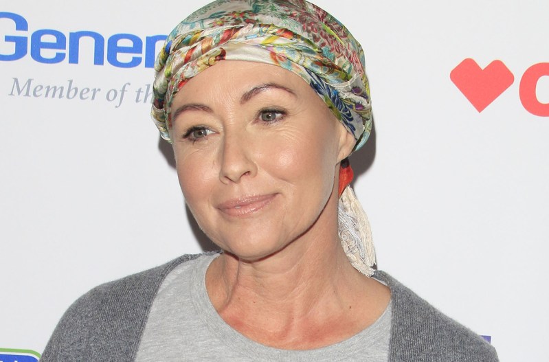 Shannen Doherty smiling at a stand up for cancer event wearing a colorful scarf on her head.