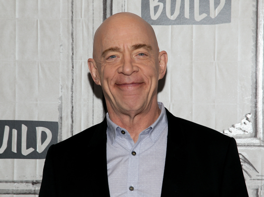 J.K. Simmons smiles at the camera wearing a blue shirt and black jacket