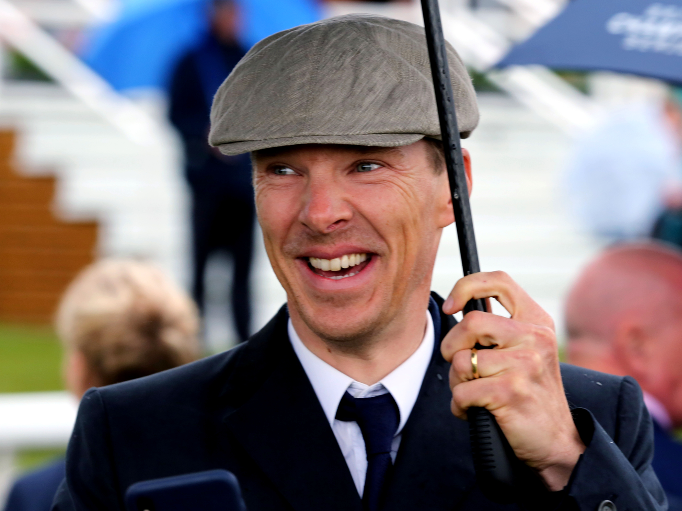Benedict Cumberbatch smiles, holding an umbrella and wearing a gray cap