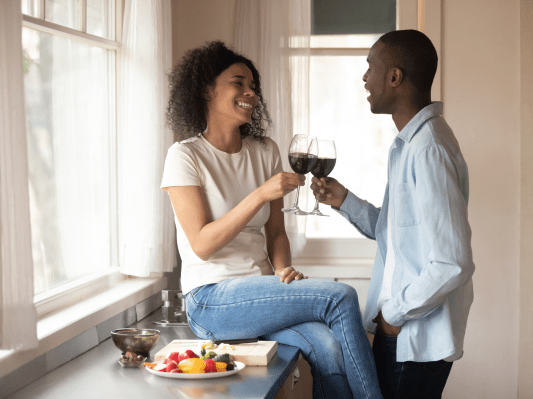 A couple sits in their kitchen, laughing, toasting with some wine