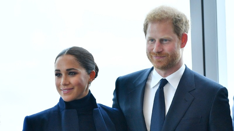 Meghan Markle, in a dark top and blue jacket, stands with Prince Harry, in a blue suit, while visiting New York
