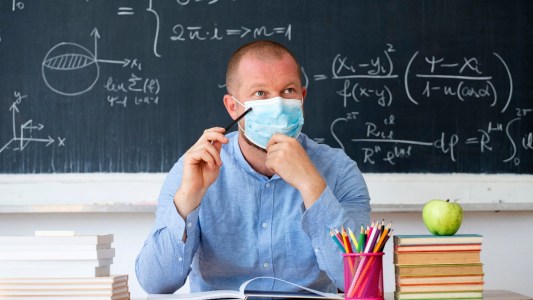 A male teacher wearing a blue shirt and a face mask sits at a desk in a classroom