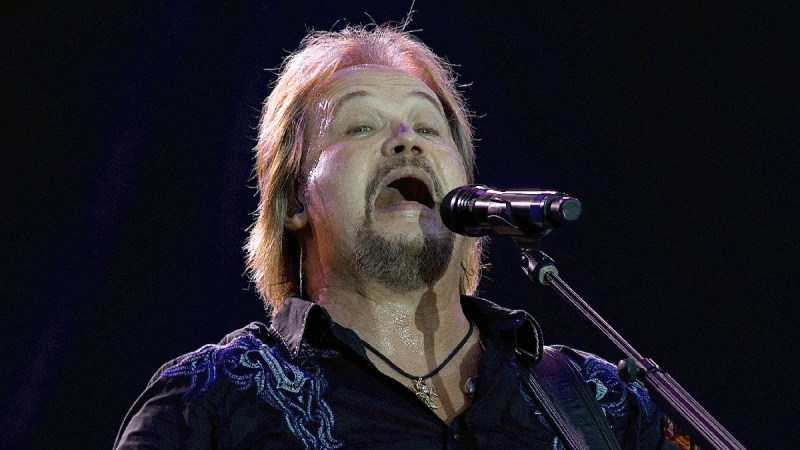 Travis Tritt wears a dark shirt and holds a guitar as he performs onstage