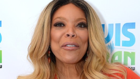 Wendy Williams wears an orange dress against a white background with blue and green letters