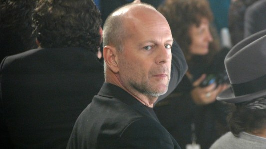 Bruce Willis wears a black suit and looks over his shoulder through the crowd at the camera