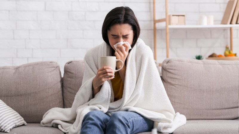 A woman bundles under a blanket as she holds a cup and blows her nose