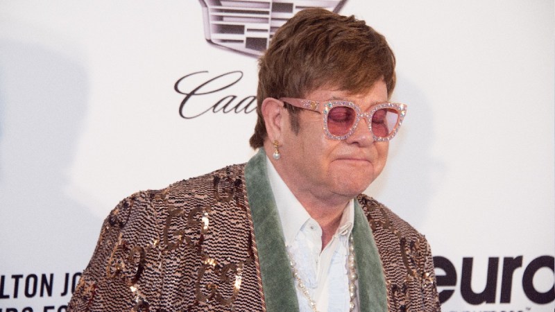 Elton John wears a bedazzled suit jacket against a white background on the red carpet