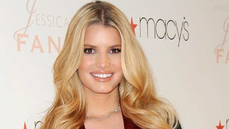 Jessica Simpson wears a red dress and black cardigan against a white background