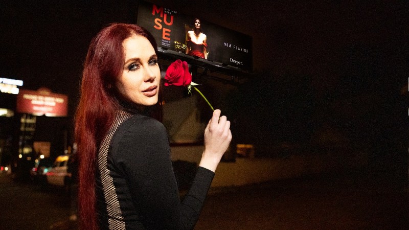 Maitland Ward wears a black dress and holds a red rose as she stands outdoors in front of a Muse 2 billboard