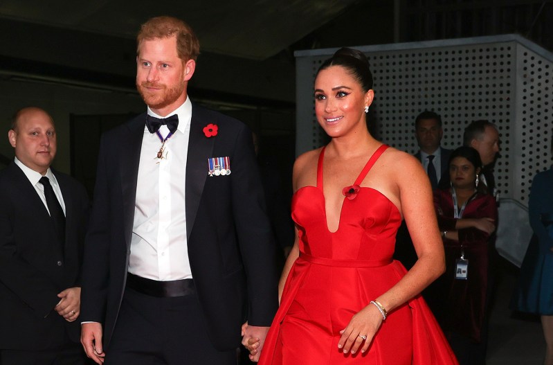 Prince Harry on the left, holding hands with Meghan Markle, both in formal wear, Markle in a red dress, Harry in a tux.