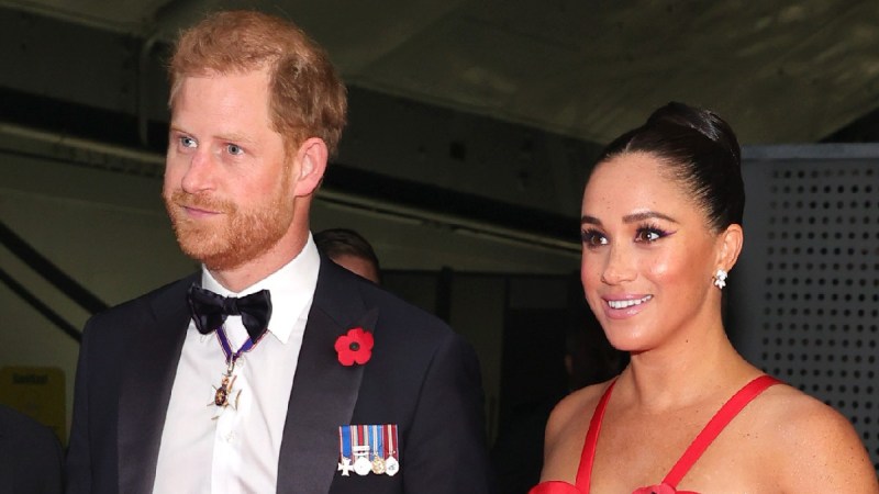 Prince Harry, in a black suit, walks the red carpet with Meghan Markle, in a red dress
