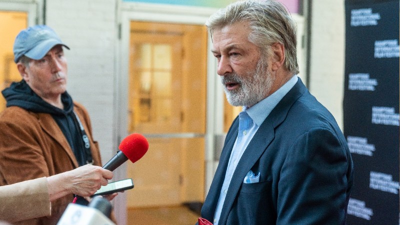 Alec Baldwin wears a blue suit as he's interviewed indoors by reporters at a film festival