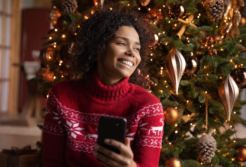 Smiling woman wearing a Christmas sweater holding a smartphone in front of a Christmas tree.
