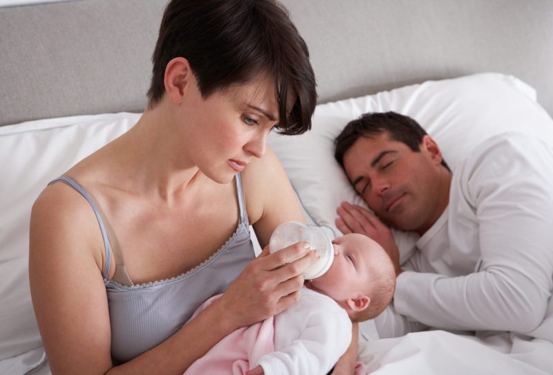 Woman feeding baby with bottle while man sleeps in bed next to her.