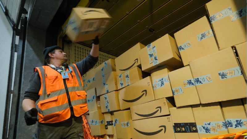 An Amazon worker loads a box into a truck while wearing an orange safety jacket