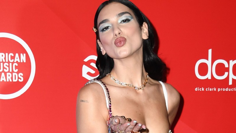 Dua Lipa blows a kiss to photographers while standing in front of a red background on the red carpet