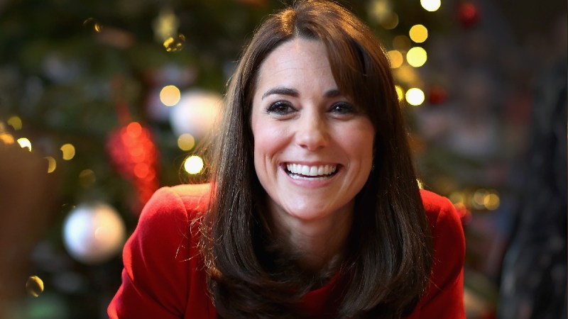 Kate Middleton, dressed in red, smiles in front of a Christmas tree