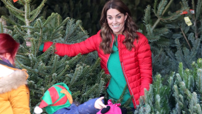 Kate Middleton wears a red jacket and green shirt while standing among Christmas trees