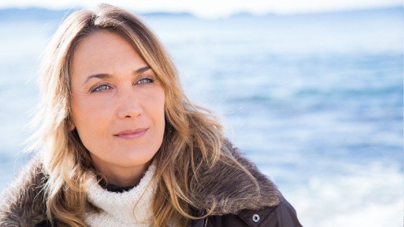A woman wears a brown coat and white sweater outdoors in front of a body of water